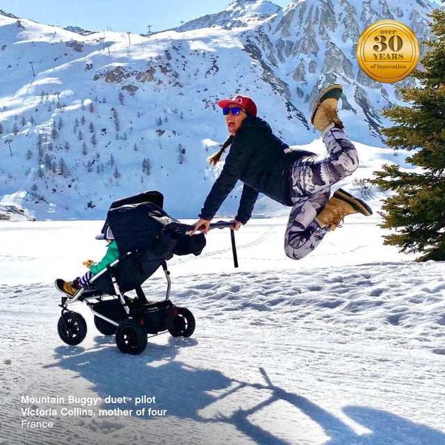 Maman active poussant la poussette duet tout en sautant de joie sur le champ de neige - Mountain Buggy duet™  pilote Victoria Collins, mère de quatre enfants, France